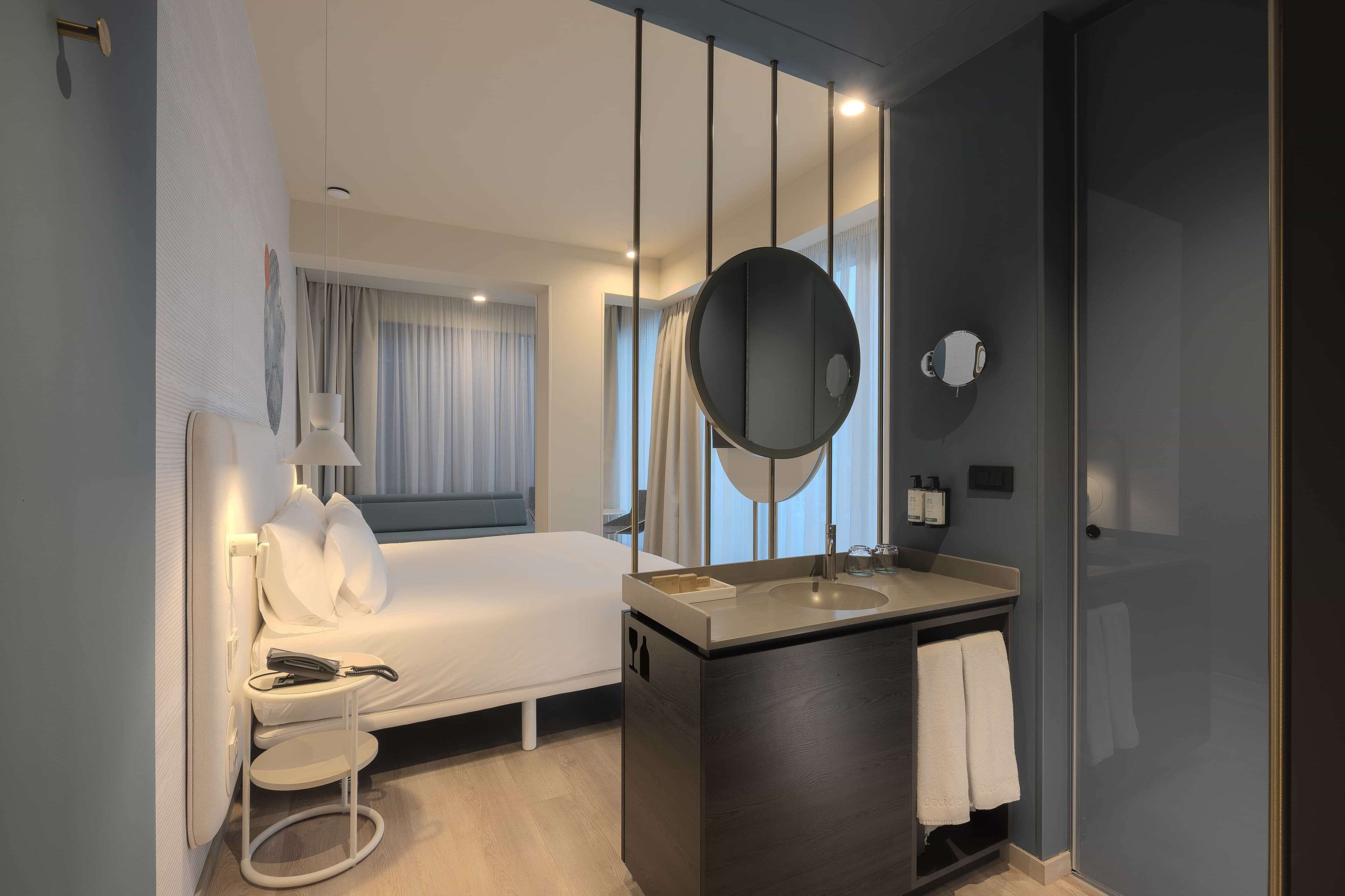 Une chambre d’hôtel moderne équipée d’un lit, d’une table de chevet, d’une lampe, d’un miroir et d’une salle de bain avec un lavabo et un porte-serviettes.