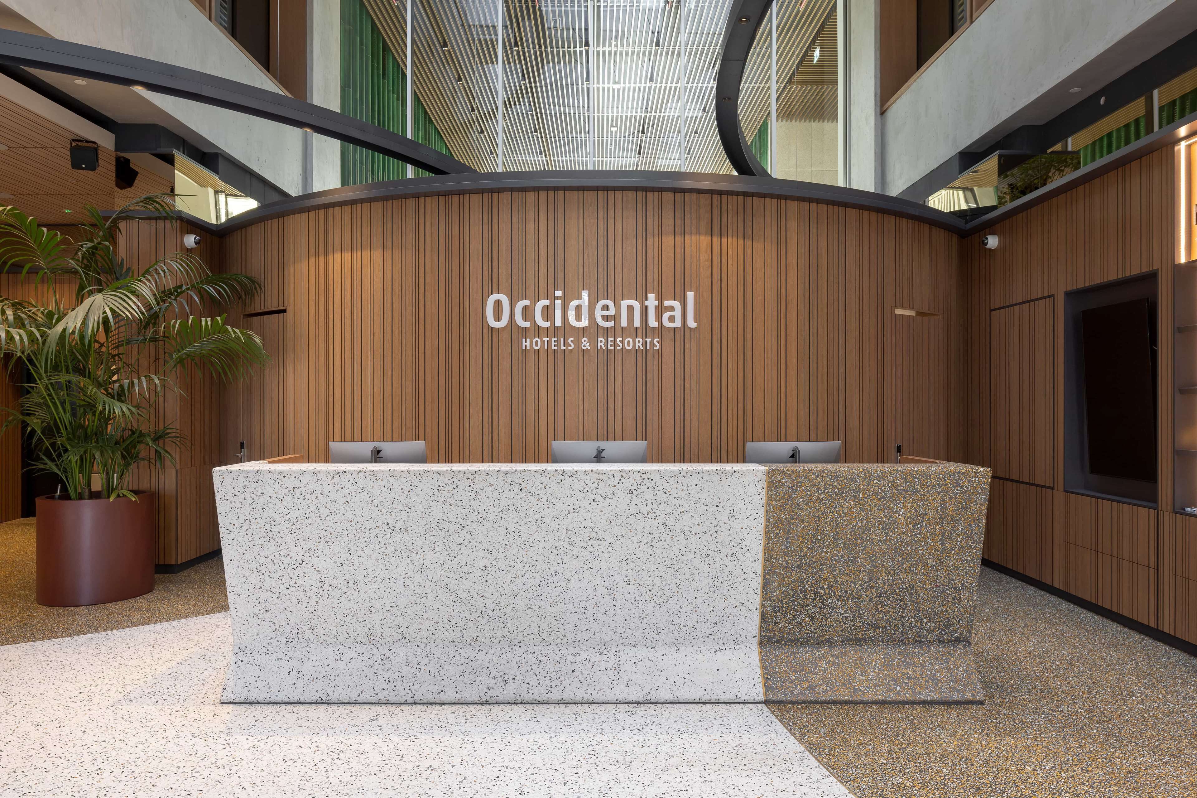 Un mostrador de recepción con un cartel que dice "Occidental Hotels & Resorts" en un vestíbulo moderno con paneles de madera y una planta grande en maceta.