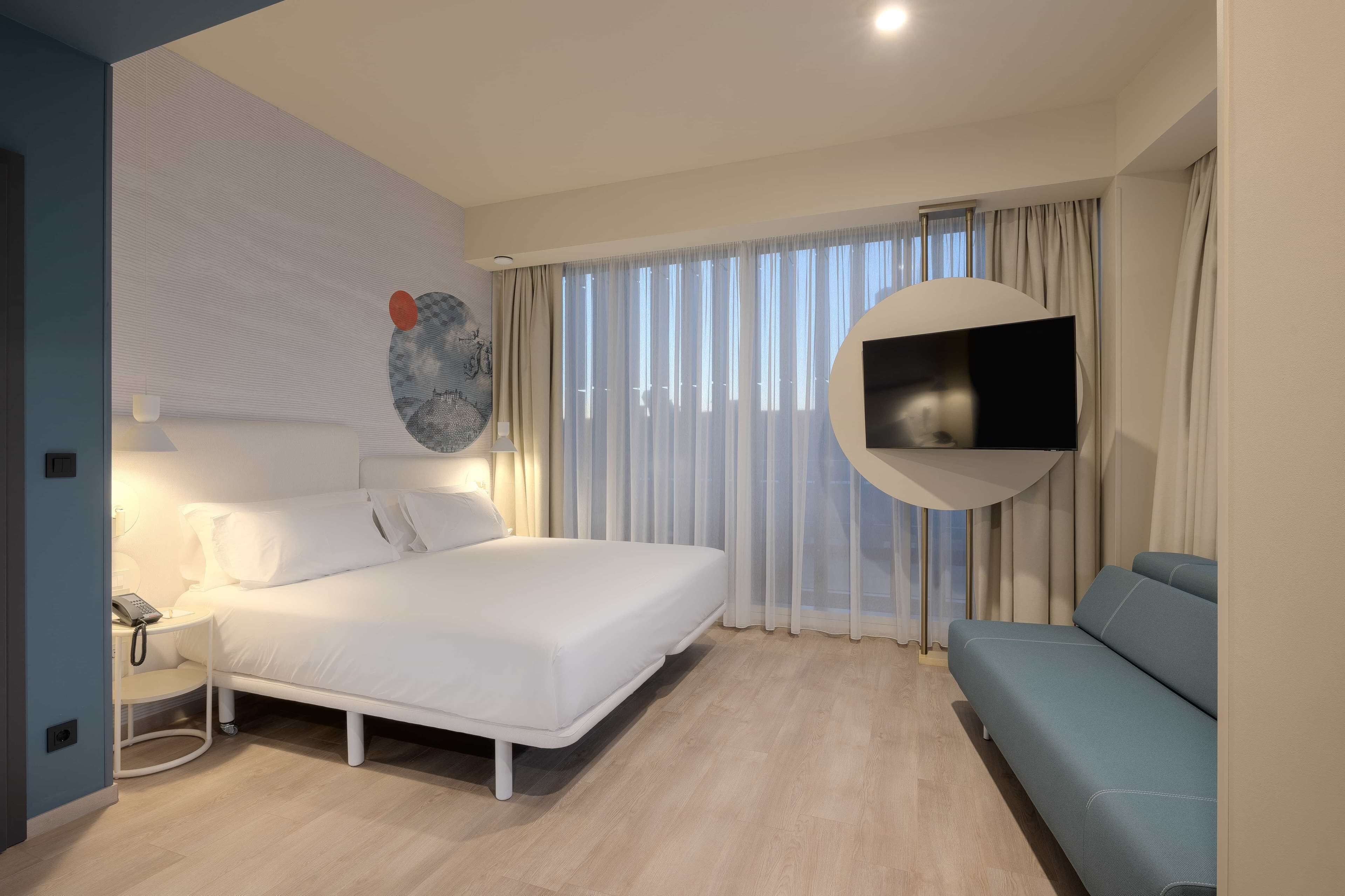 A modern hotel room with a bed, a TV mounted on the wall, a sofa, and a bedside table with a lamp.