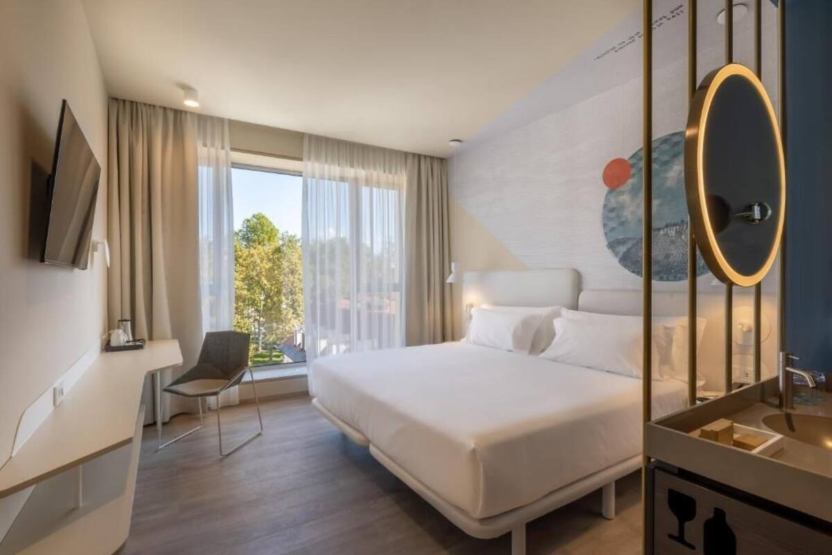 Ein Hotelzimmer mit Bett, Schreibtisch, Stuhl, Fernseher und einem großen Fenster mit Vorhängen.