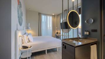 A modern hotel room with a large bed, a bedside table with a lamp, a large mirror, and a bathroom with a sink and mirror.