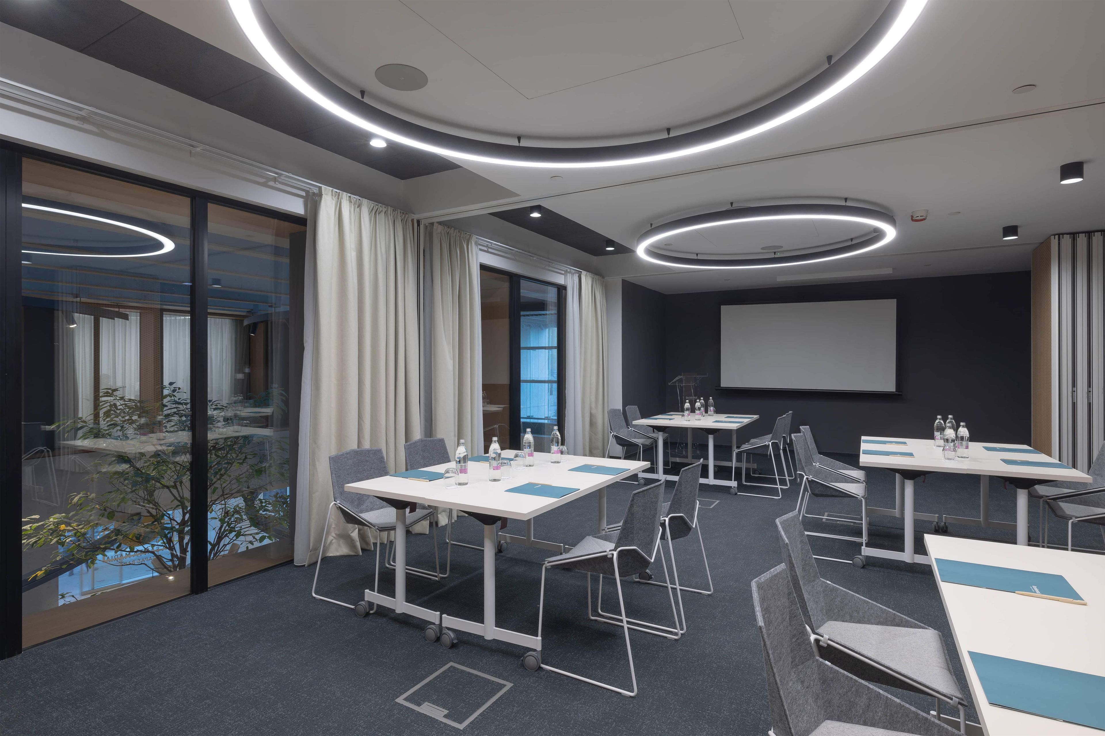 A conference room with round tables, chairs, a projector screen, and a large window with curtains.
