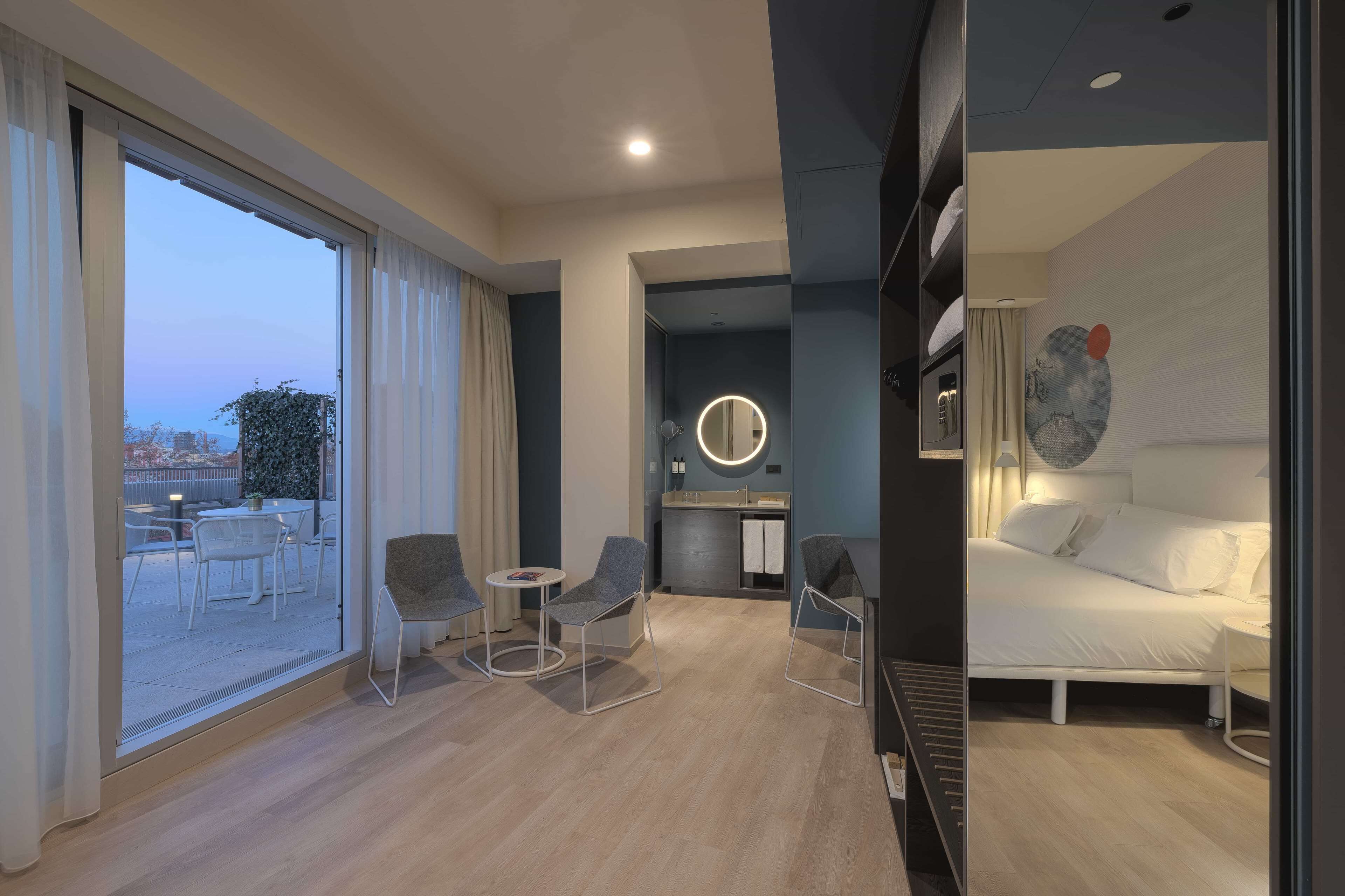 Una habitación de hotel moderna con un ventanal grande, una cama, una mesita redonda pequeña y dos sillas.