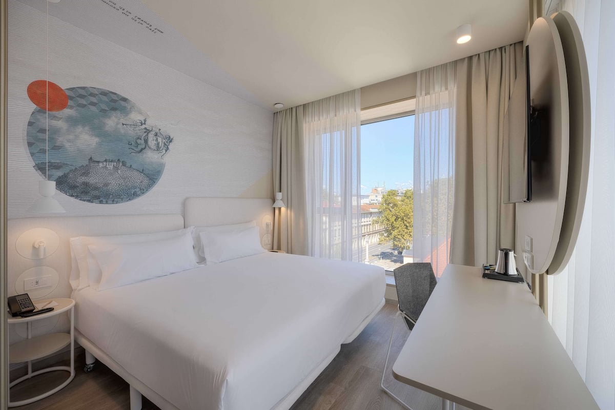 Ein Hotelzimmer mit einem großen Bett, einem Schreibtisch, einem Stuhl, einem Fernseher und einem Vorhangfenster.