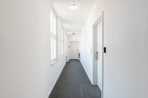 A narrow, well-lit hallway with white walls and a grey carpet. There are windows on the left side, a fire extinguisher on the right, and a door at the end.