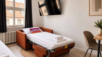 Ein kleines Hotelzimmer mit einem Einzelbett, einem an der Wand montierten Fernseher, einem Tisch mit einer Pflanze und Blick auf Gebäude durch das Fenster.