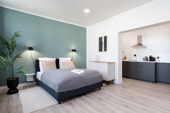 A modern bedroom with a bed, bedside lamps, a nightstand, a potted plant, and a view into a kitchen area. - nestup Hildesheim Zentral (Hildesheim)