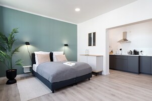 Un dormitorio moderno con cama, lámparas de noche, mesita de noche, una planta en maceta y vistas a una zona de cocina.