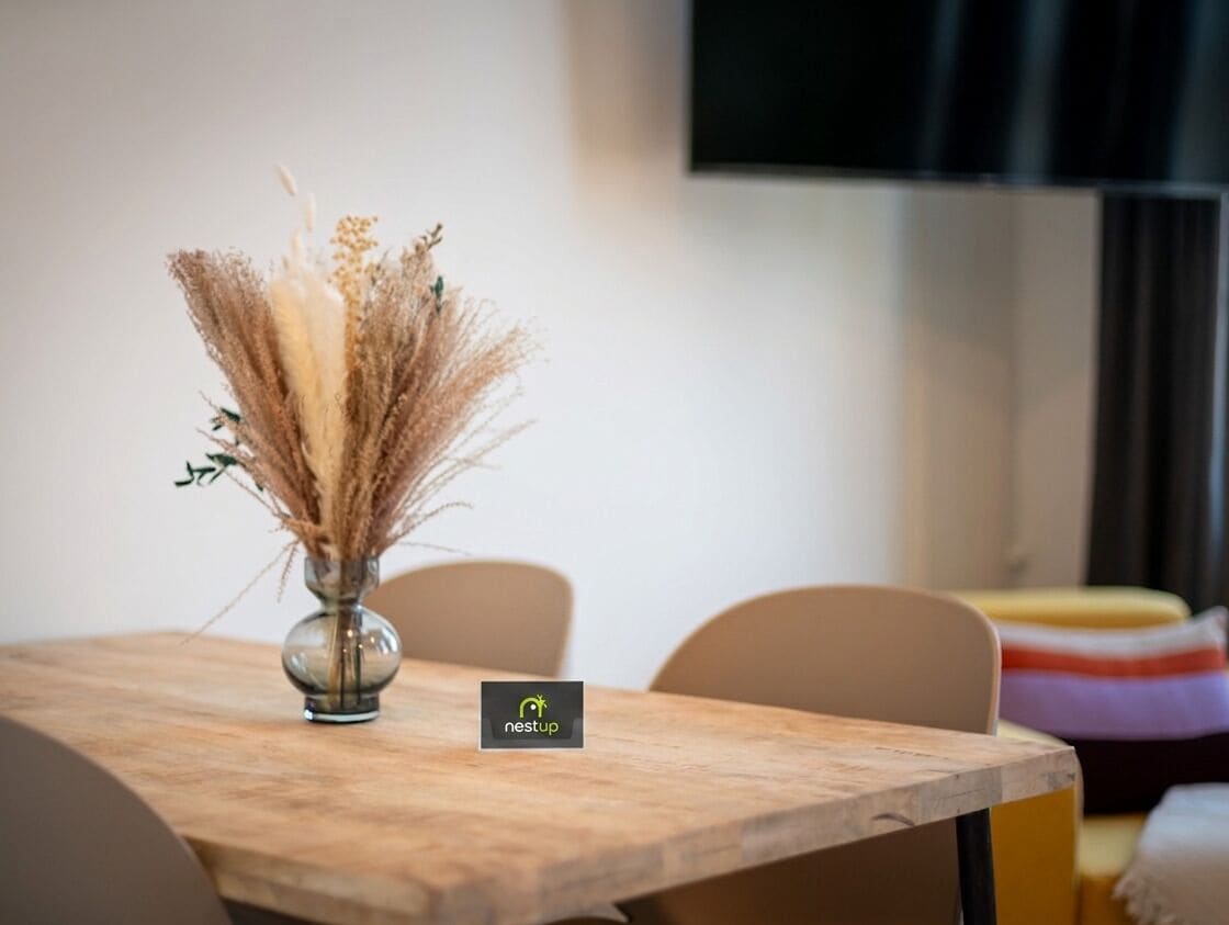Et bord med en vase med tørrede planter, en stol og en uskarpt baggrund med et fjernsyn.