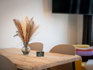 A table with a vase containing dried plants, a chair, and a blurred background with a television. - nestup Hildesheim Zentral (Hildesheim)