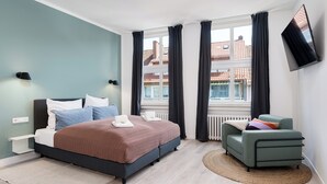 A modern bedroom with a bed, bedside tables, a TV, and a view of buildings. - nestup Hildesheim Zentral (Hildesheim)