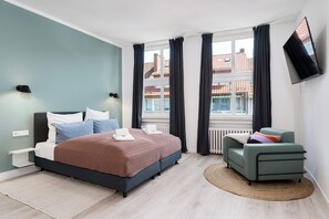 A modern bedroom with a bed, bedside tables, a TV, and a view of buildings. - nestup Hildesheim Zentral (Hildesheim)
