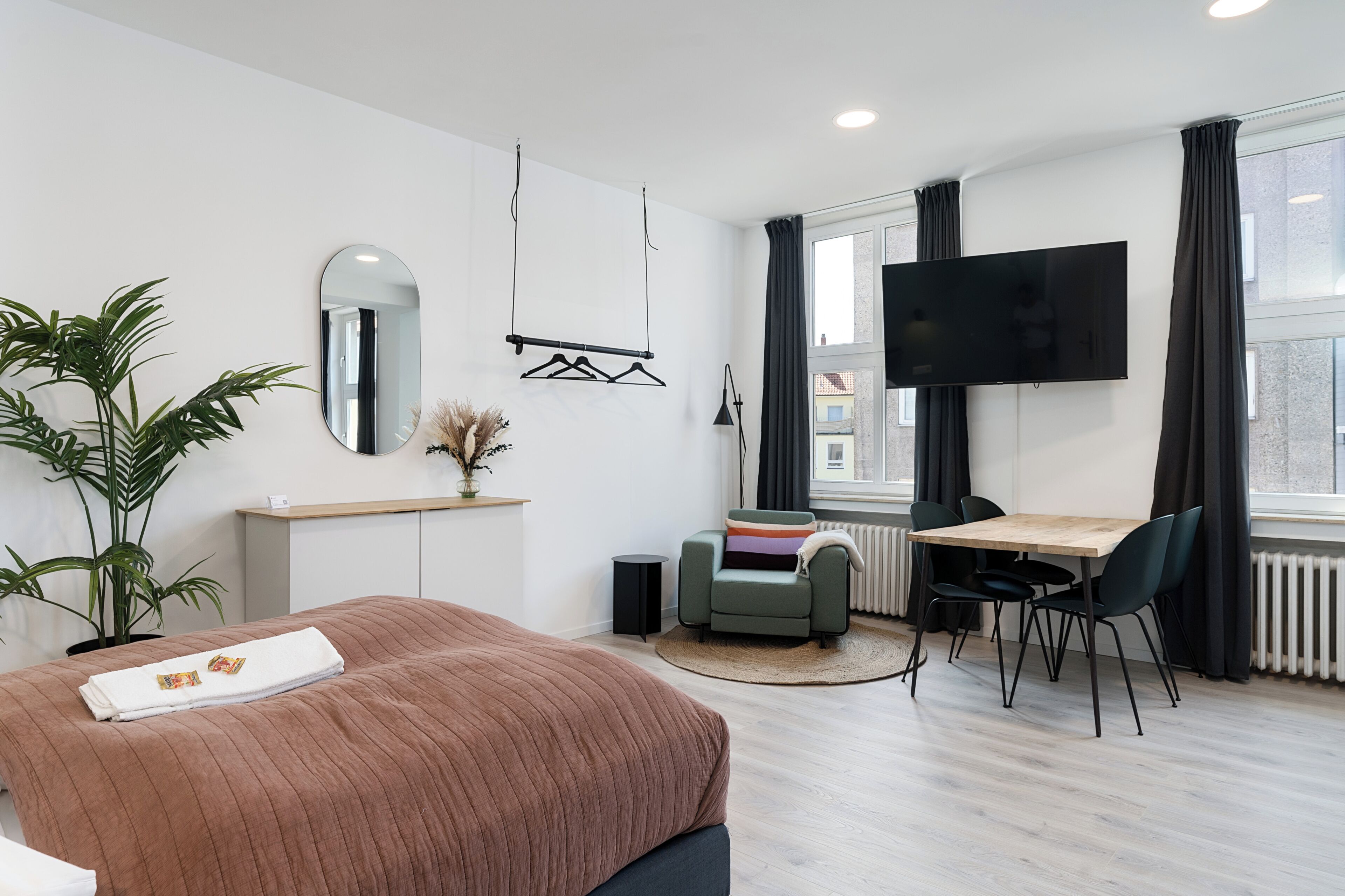 A modern hotel room with a bed, a desk, a chair, a TV, a plant, and a window with curtains.