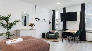 A modern hotel room with a bed, a desk, a chair, a TV, a plant, and a window with curtains.