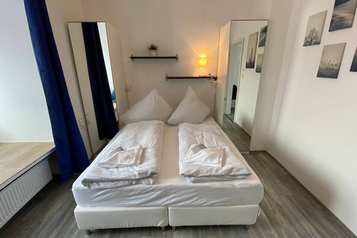 A double bed with white linens and pillows, a bedside table with a lamp, a small shelf with a plant, and a mirror on the wall.
