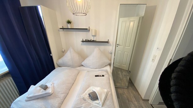 Una cama doble con toallas, una mesita de noche, un estante en la pared con una planta y una puerta a la derecha.