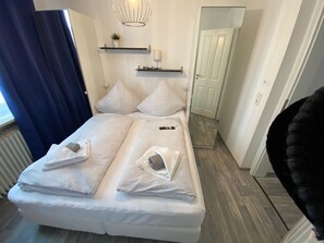 Un lit double avec des serviettes, une table de chevet, une étagère murale avec une plante et une porte sur le côté droit.