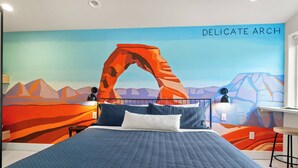 A bedroom with a bed, a desk, and a chair. The wall features a painting of a mountain range and a large arch. - Downtown Moab • Hot Tub • Walk to Dining & Trails (Moab)