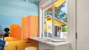 Una stanza con un murale colorato, una finestra vista su un edificio giallo, due sgabelli e un tavolo con dei documenti.