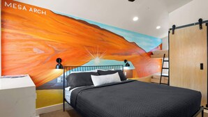 A bedroom with a mural of a sunset, a bed with black bedding, a desk, and a wardrobe.