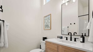 A modern bathroom with a white toilet, wooden vanity, and a large mirror with lights.