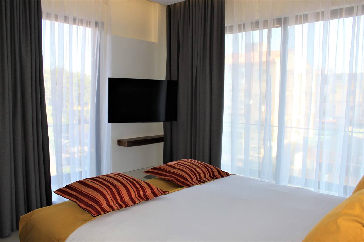 A hotel room with a bed, two pillows, a flat-screen TV mounted on the wall, and large windows with sheer curtains.
