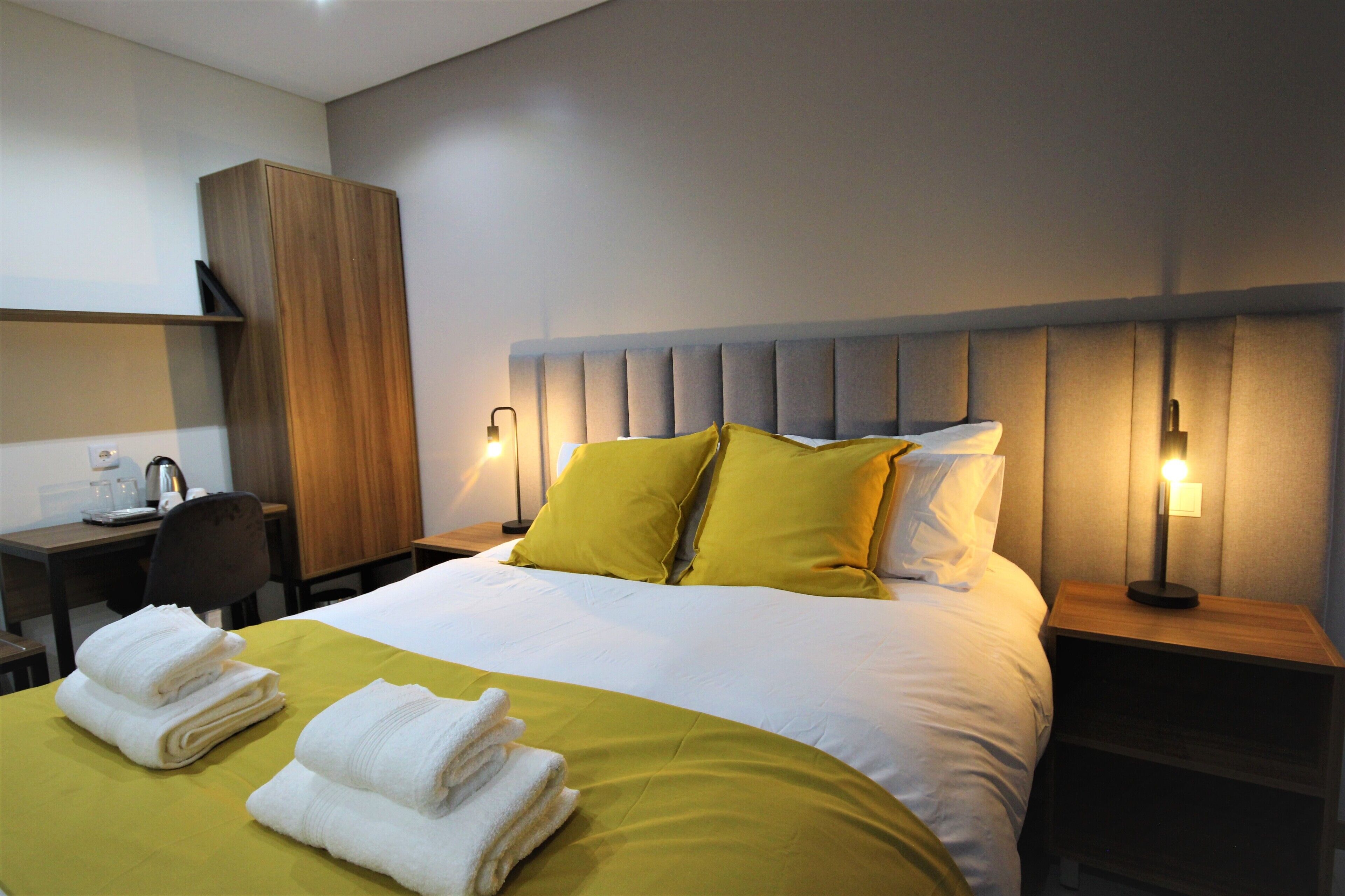A hotel room with a neatly made bed, a desk with a chair, and a nightstand with a lamp.