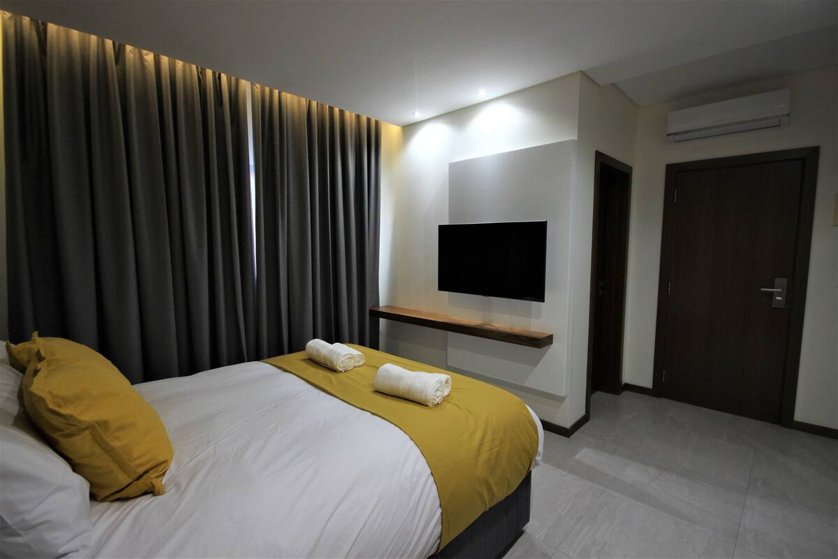 A hotel room with a bed, a TV mounted on the wall, a door, and a window with curtains.