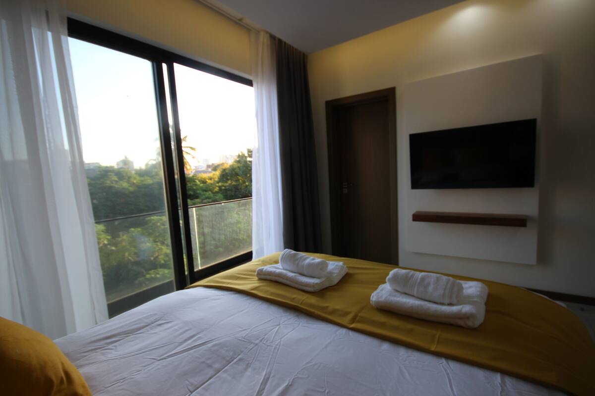 A hotel room with a large bed, a TV mounted on the wall, and a balcony with a view of greenery.