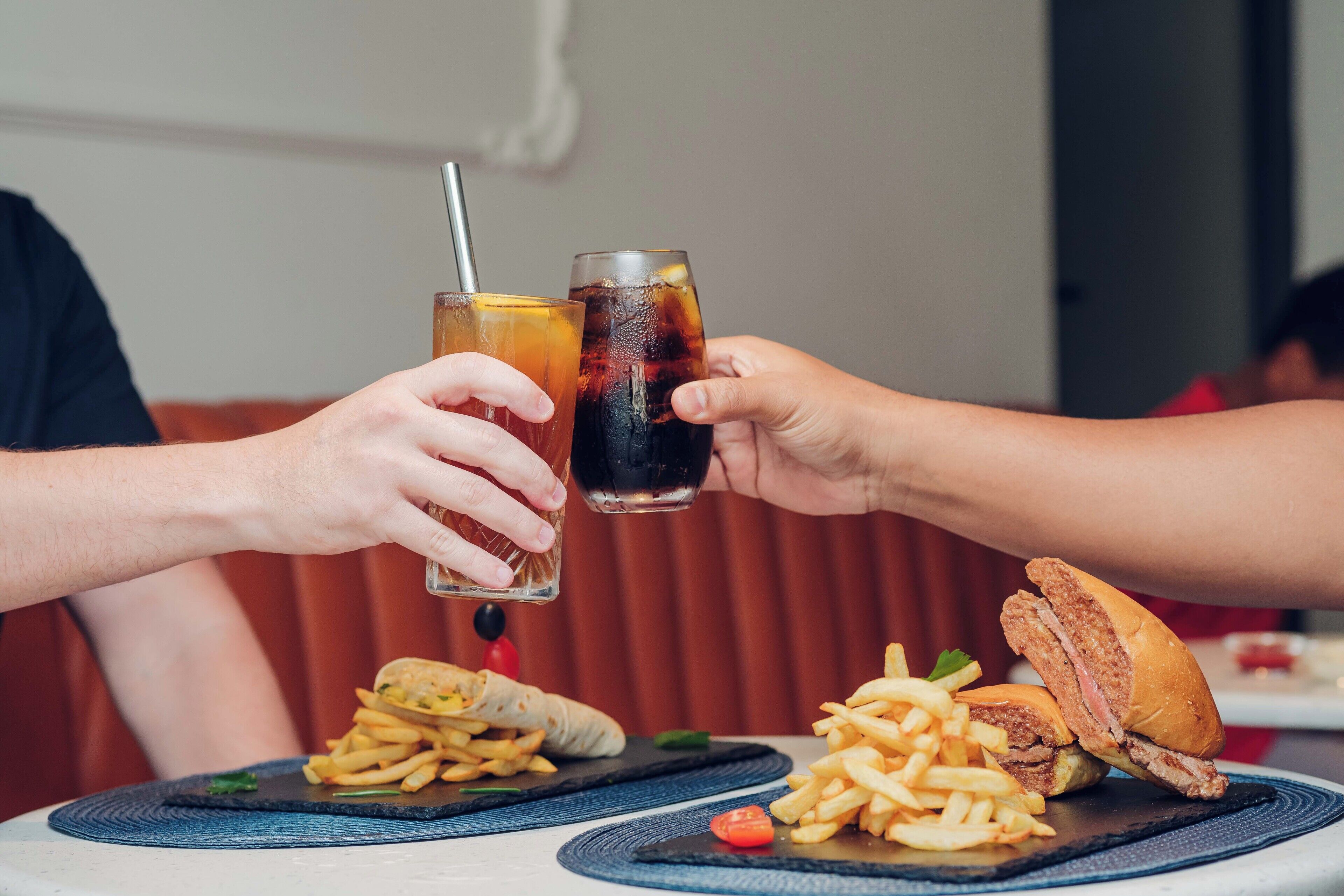 Une personne tenant deux verres d’une boisson sombre, probablement du cola, avec une paille, et une assiette contenant un sandwich et des frites.