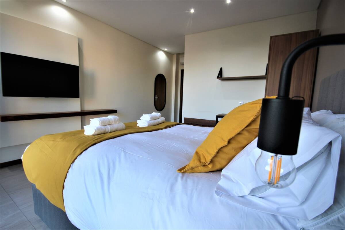 A modern hotel room with a neatly made bed, a mounted TV, and a unique ceiling light fixture.