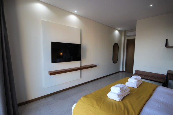 Quarto de hotel moderno com uma cama grande, televisão de tela plana na parede e um banco de madeira junto à porta.