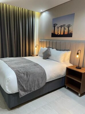 A modern bedroom with a large bed, grey bedding, a grey throw blanket, a headboard with built-in lighting, a nightstand with a lamp, and a painting of trees on the wall.