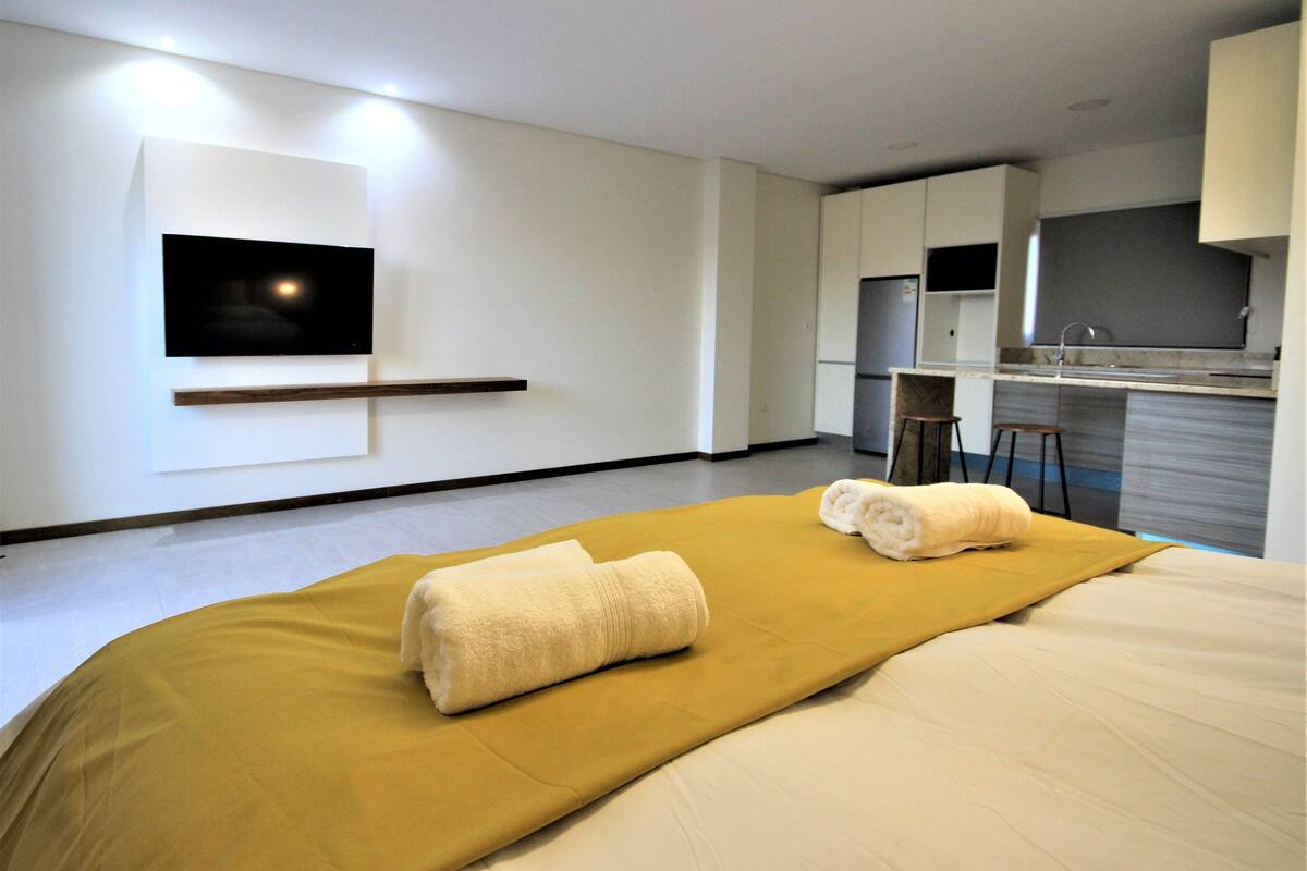 A modern hotel room with a bed, a flat-screen TV mounted on the wall, a kitchen area with a refrigerator, and a small bar stool.