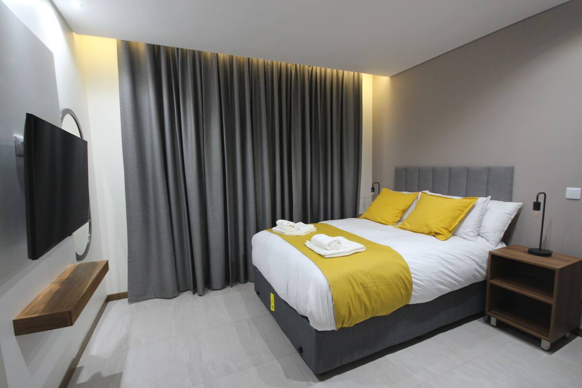 A modern hotel room with a large bed, a flat-screen TV, a bedside table, and a built-in shelf.