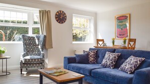 A living room with a blue sofa, patterned armchair, and a wooden coffee table. A clock and a framed picture of a soup can are on the wall.