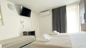 A hotel room with two beds, a flat-screen TV, an air conditioning unit, and a window with curtains. - bed and book (Rimini)