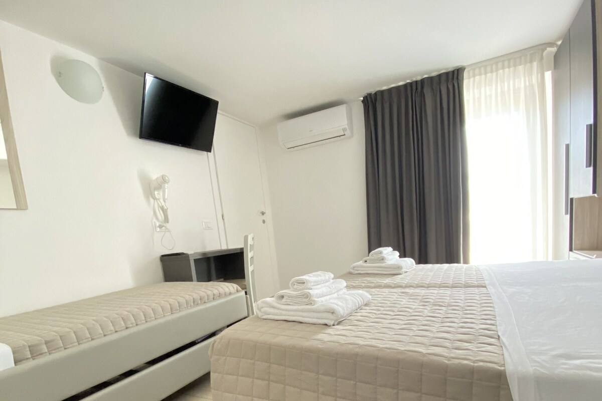 A hotel room with two beds, a flat-screen TV, an air conditioning unit, and a window with curtains.