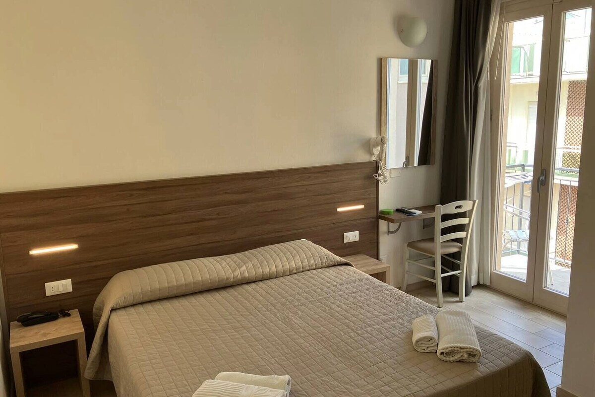 A hotel room with a bed, a desk, a chair, a mirror, and a view of the outside balcony.