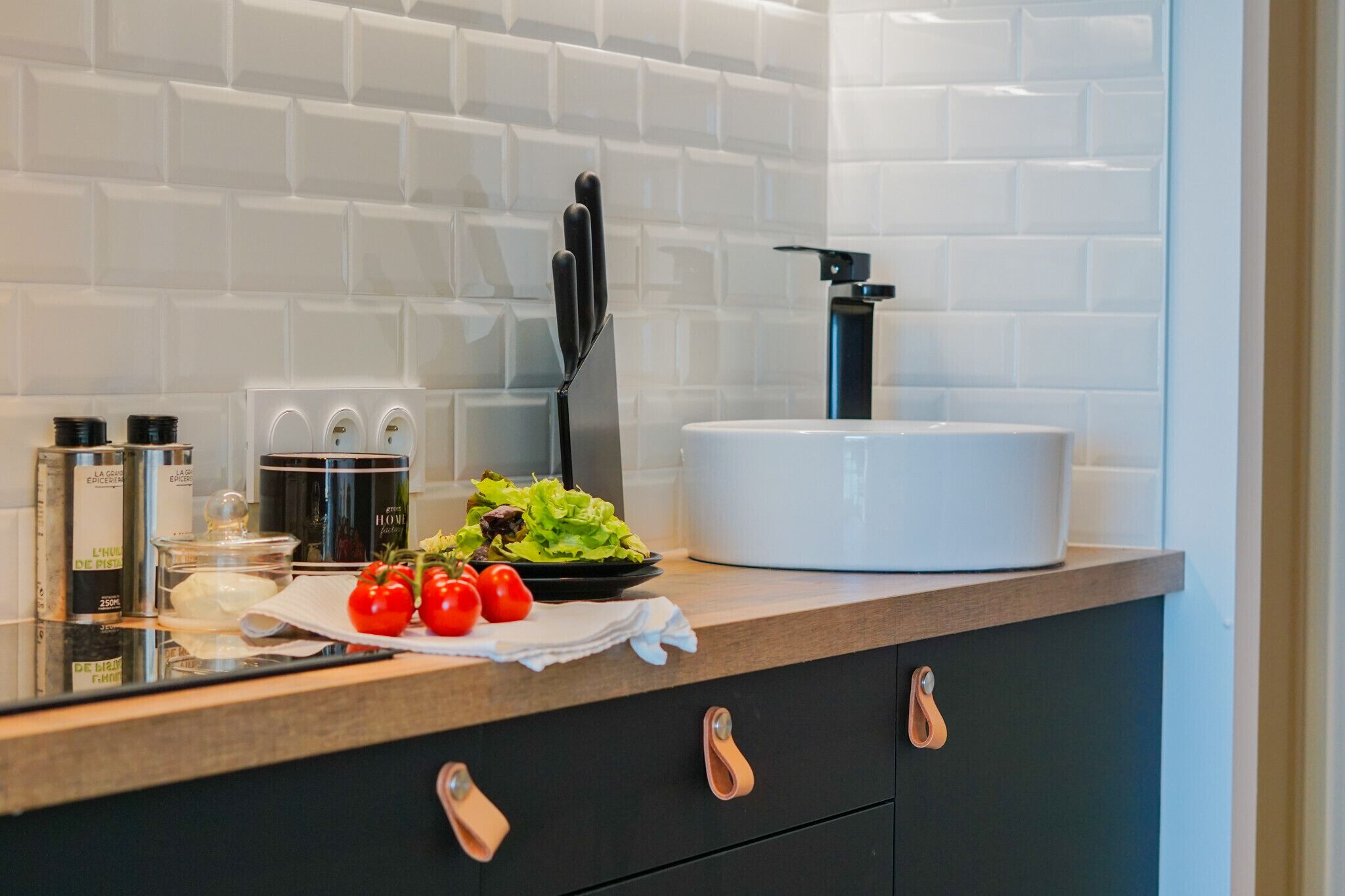 Uma cozinha moderna com painel de azulejos branco, um conjunto de facas preto, um cozido lento branco e tomates frescos na bancada.