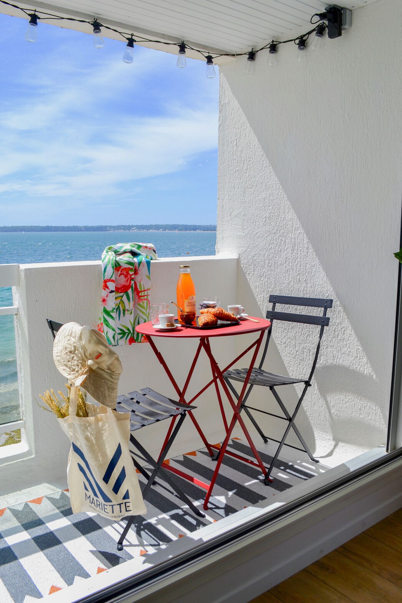 A balcony with a table and chair, a bottle of orange juice, and a bag with the Mariette logo.