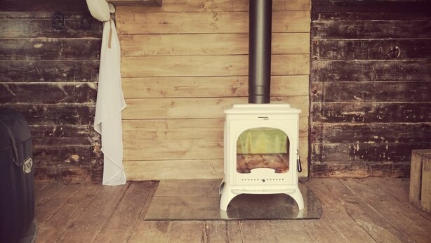 Une pièce rustique en bois, avec un poêle à bois, un rideau blanc et une petite étagère où sont empilés des bûches.