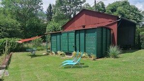 Un abri de jardin en métal vert, avec un toit rouge, entouré d’une cour verdoyante où se trouvent deux chaises de jardin bleues et un parasol rouge.