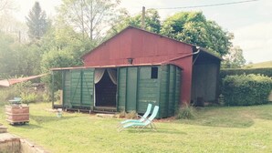 Une grange rouge à l’aspect rustique, avec une porte verte, un petit porche et une chaise de jardin bleue.