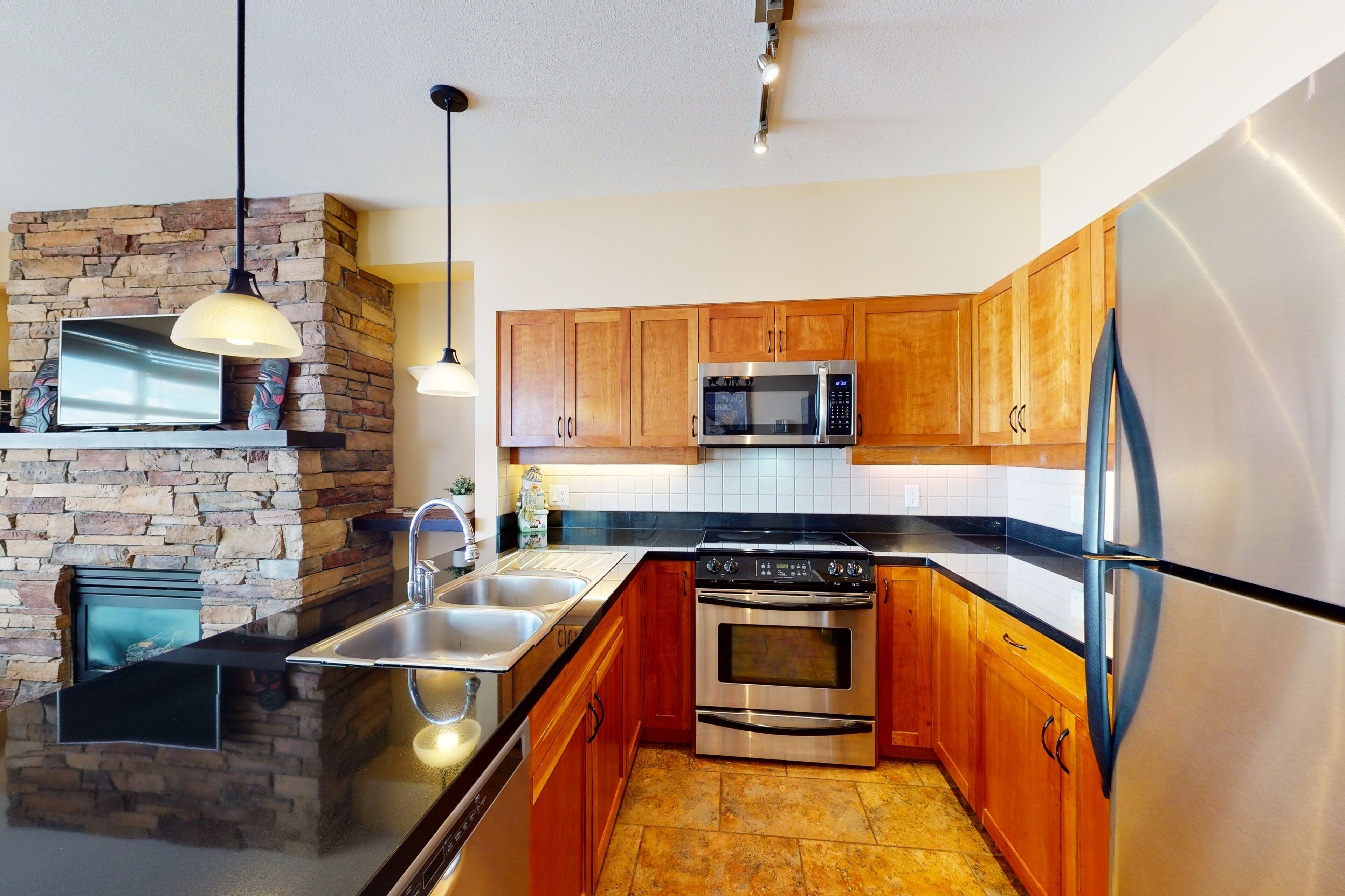 A modern kitchen with a stone fireplace, stainless steel appliances, wooden cabinets, and pendant lighting.