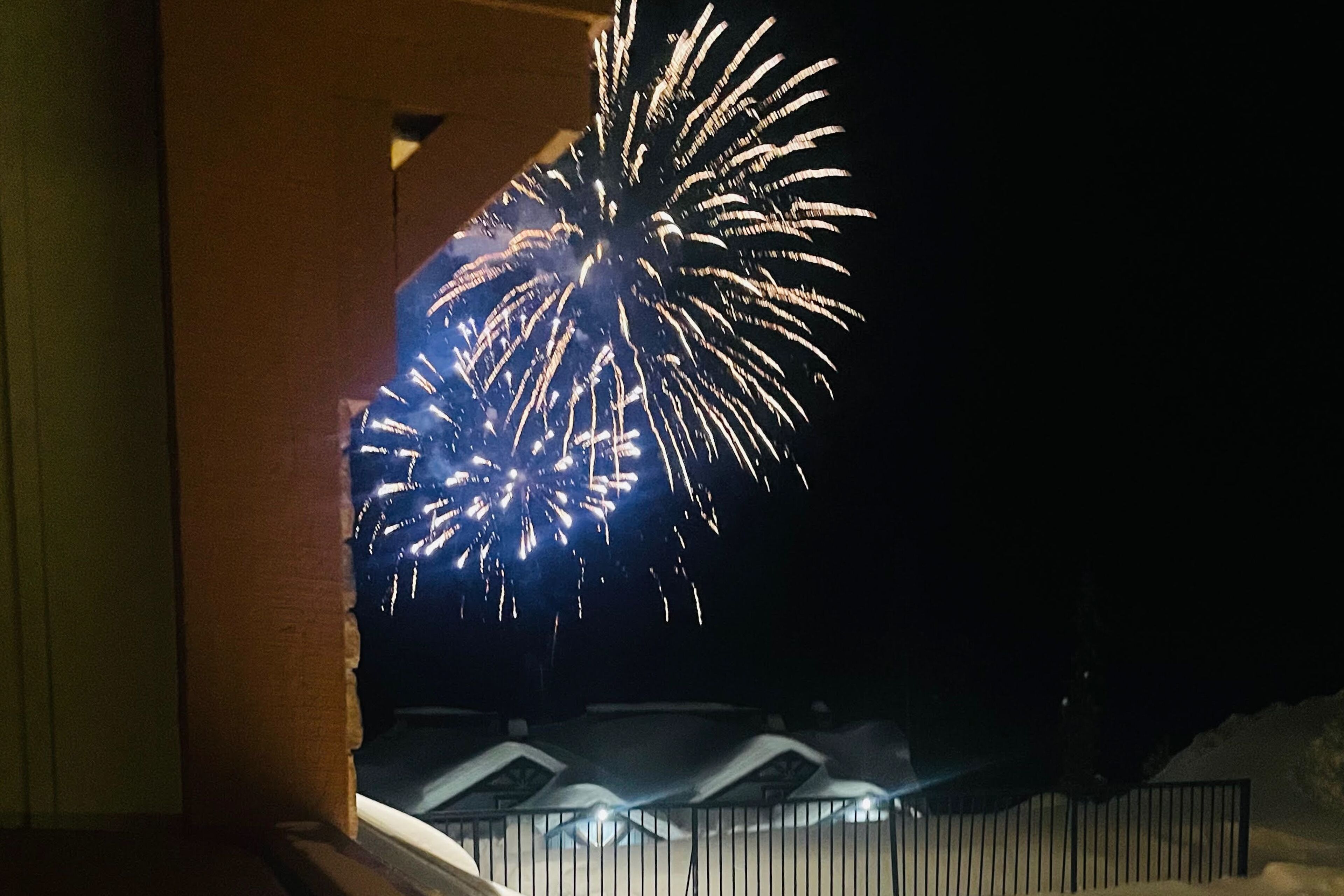 A nighttime view of fireworks with a building and a fence in the foreground.