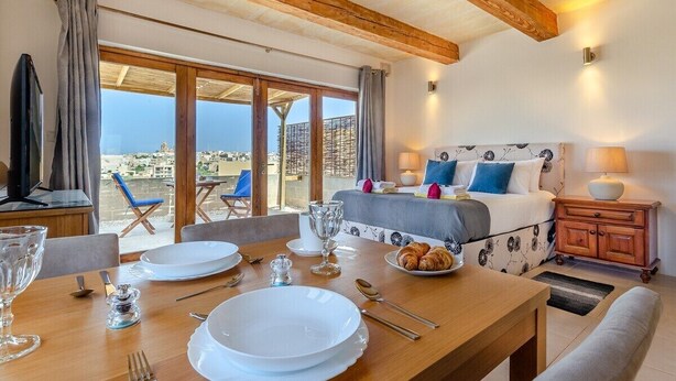 Een hotelkamer met balkon, een bed met bloemenmotief hoofdbord, een eettafel gedekt met borden en glazen, en uitzicht op de stad.