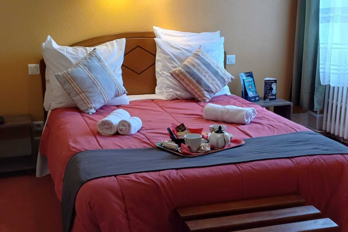 A neatly made bed with a red bedspread, pillows, and rolled towels. A tray with tea and snacks is placed on the bed.