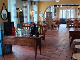Um restaurante com mesas e cadeiras de madeira, um espelho grande na parede e vista para a rua através de janelas amplas.