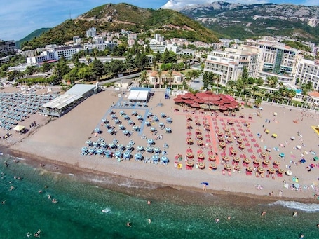 An aerial view of a beach with rows of sun loungers and umbrellas, a hotel in the background, and clear blue waters.